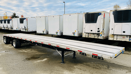 A white flatbed trailer in a cement lot surrounded by parked semi trailers.