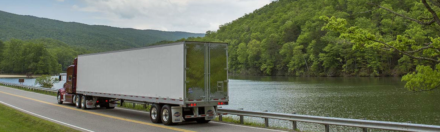 A semi truck pulling a trailer and driving along a river that's bordered by forest.